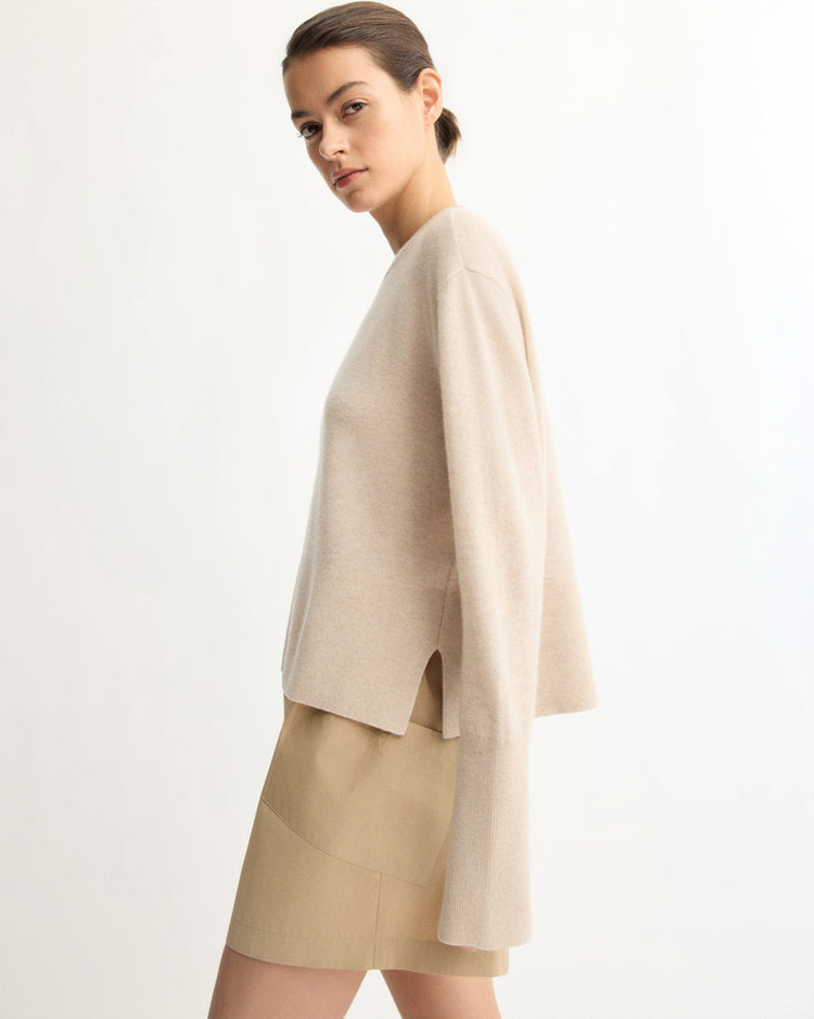 A woman stands sideways against a plain white background, wearing the Arlo Mott Warren Classic Crew Neck sweater in beige with side slits and a light tan mini skirt.