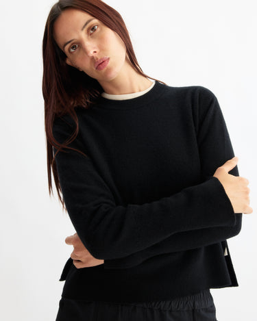 A woman with long brown hair, wearing the Arlo Mott Warren Classic Crew Neck in Black and black pants, stands against a plain white background with her arms crossed and head slightly tilted.