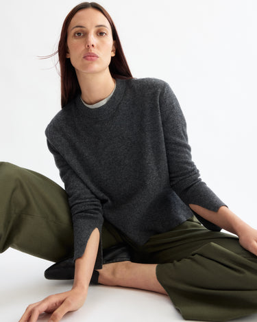 A woman with straight brown hair sits on the floor against a white background, wearing an Arlo Mott Warren Classic Crew Neck in Charcoal and olive green wide-leg pants, her expression calm and neutral.