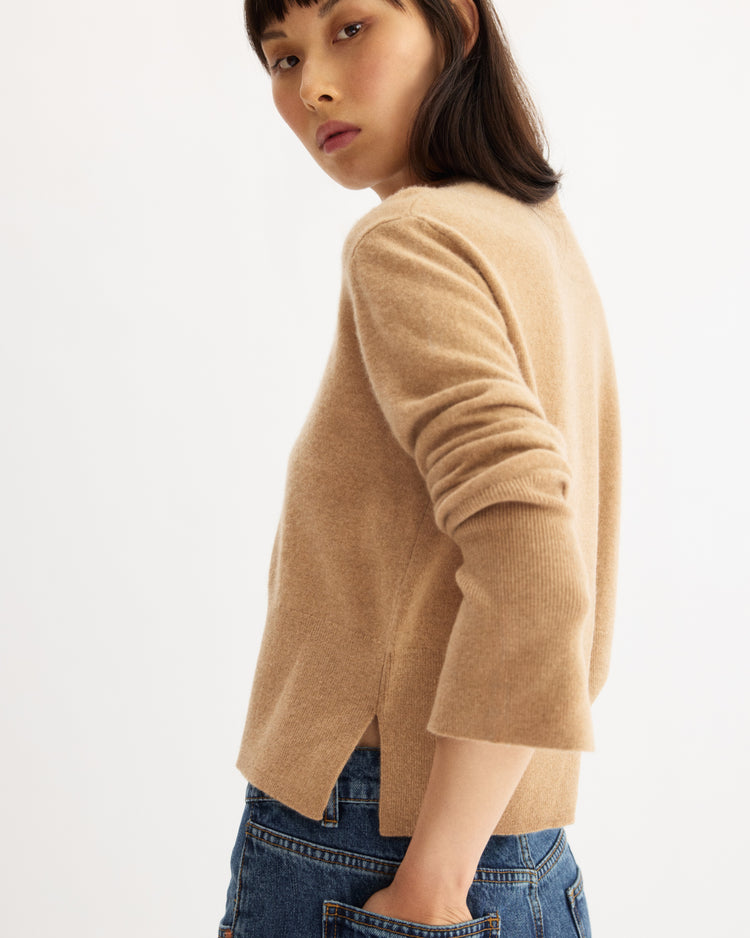 A person with straight dark hair wears the Arlo Mott Warren Classic Crew Neck in Honey and blue jeans, standing against a plain light background and looking over their shoulder with a neutral expression.