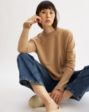 A person with short, straight hair wears the Arlo Mott Warren Classic Crew Neck in Honey, wide-leg blue jeans, and beige shoes while sitting on the floor against a plain white background, resting their head on one hand.
