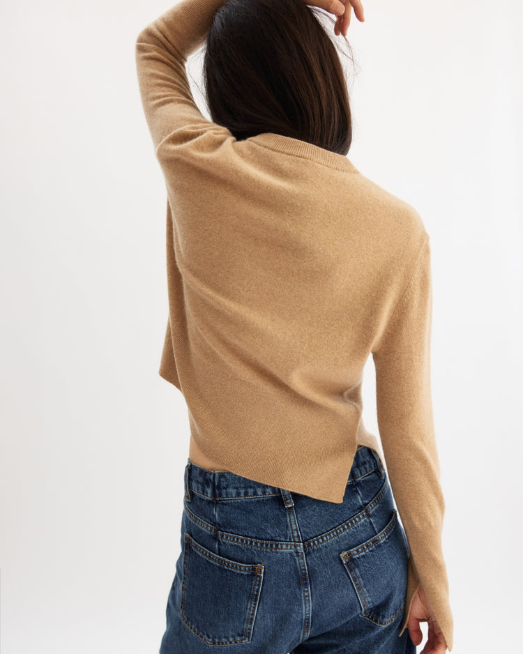 A person with long dark hair, wearing an Arlo Mott Warren Classic Crew Neck in Honey and blue jeans, stands with their back to the camera and one arm raised to touch their head. The background is plain white.