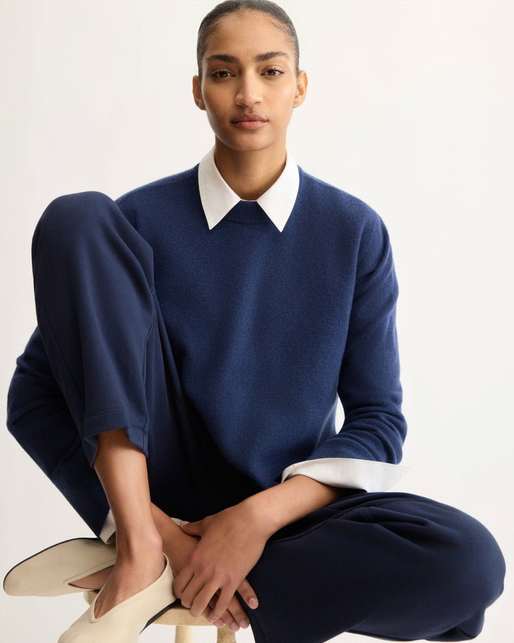 A person with tied-back hair sits on a stool, wearing the Arlo Mott Warren Classic Crew Neck in Navy over a white collared shirt, navy pants, and white slip-on shoes, with an arm resting on their knee against a plain light background.