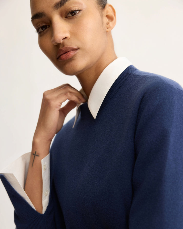 A person with short hair wears the Arlo Mott Warren Classic Crew Neck in Navy over a white collared shirt. They look at the camera with a neutral expression, resting one hand near their chin and revealing a small cross tattoo on their wrist.