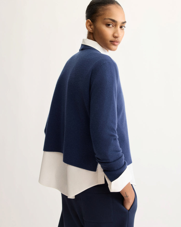 A woman seen from behind, glancing over her shoulder, wears the Arlo Mott Warren Classic Crew Neck in Navy over a white collared shirt with navy trousers, set against a light grey background.