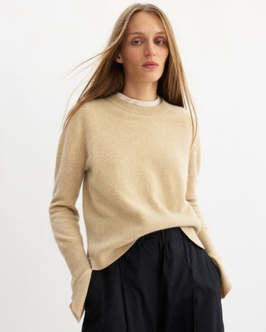 A person with long, straight blonde hair wears the Arlo Mott Warren Classic Crew Neck in Oatmeal over a white top and loose black pants, standing against a plain white background with a neutral expression.