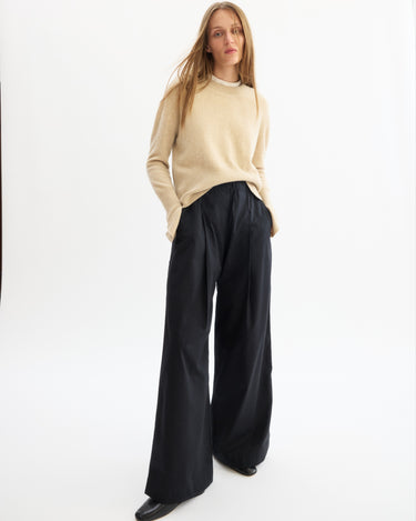 A woman stands against a white background, wearing the Arlo Mott Warren Classic Crew Neck in Oatmeal, loose black wide-leg trousers, and black shoes. Her hands are in her pockets and her straight blonde hair is worn loose.