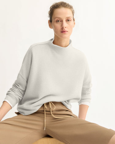 A person with light skin and short, curly hair models the Arlo Mott Penn Roll Neck Top in Grey, paired with tan pants, while seated casually on a wooden object against a plain white background.