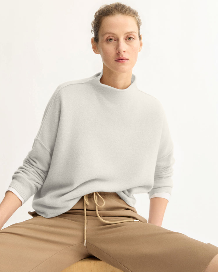 A person with light skin and short, curly hair models the Arlo Mott Penn Roll Neck Top in Grey, paired with tan pants, while seated casually on a wooden object against a plain white background.