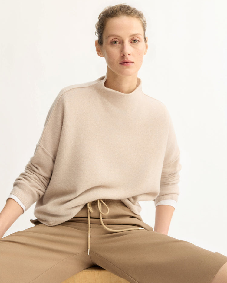 A person with short, curly hair wears the Arlo Mott Penn Roll Neck Top in Beige and tan pants, seated on a wooden object against a plain light background. Their expression is neutral and relaxed.