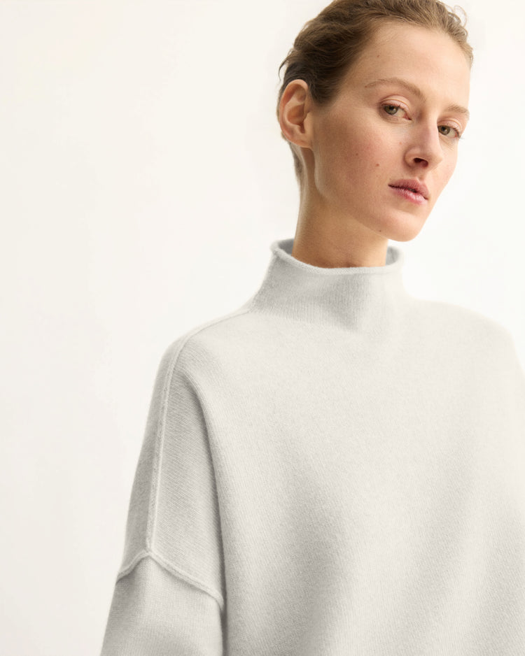 A woman with light skin and short, pulled-back hair wears the Arlo Mott Penn Roll Neck Top in Grey. She gazes slightly to the side with a neutral expression, standing against a plain, light background.