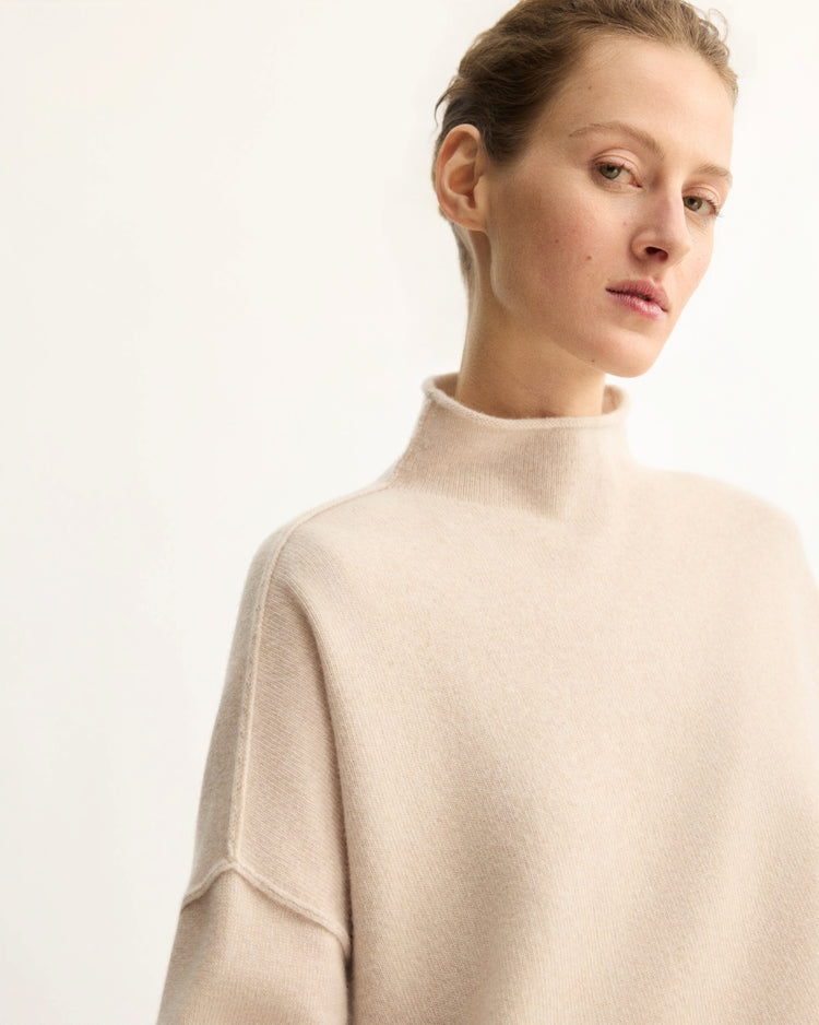 A person with short, pulled-back hair wears the Arlo Mott Penn Roll Neck Top in Beige, looking slightly to the side against a plain light background.