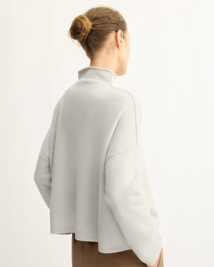 Shown from behind against a plain background, a person with light brown hair in a bun wears the Arlo Mott Penn Roll Neck Top in Grey paired with brown pants.