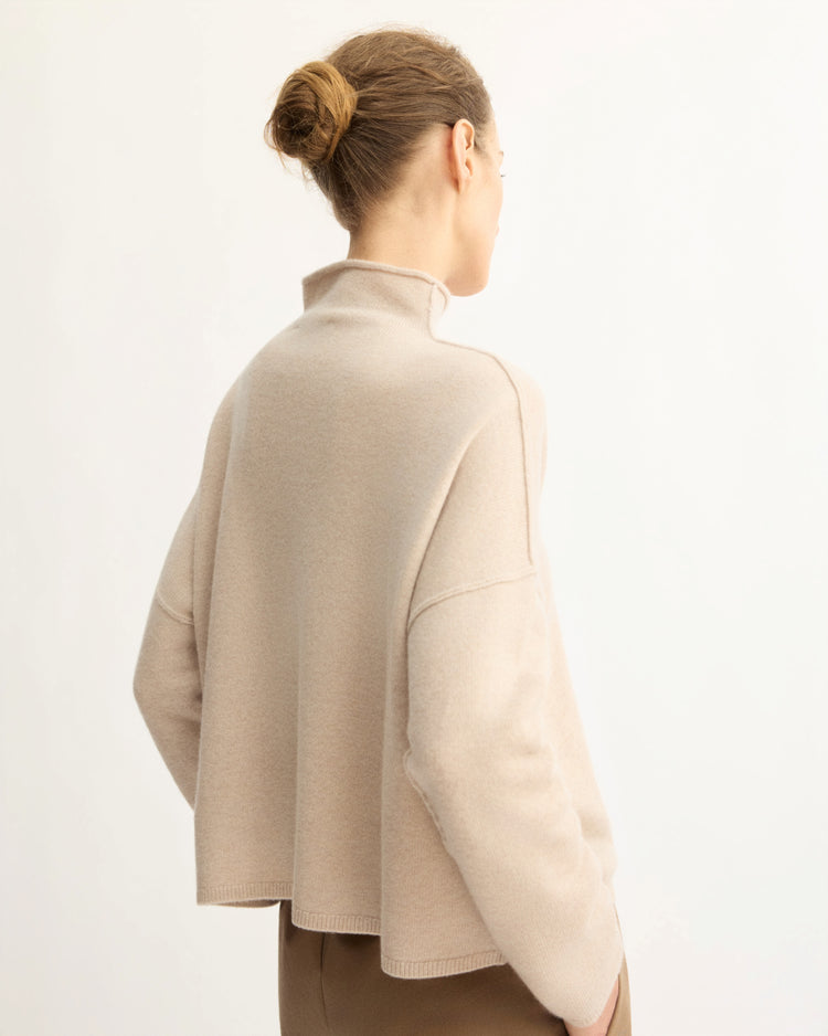 A person with light brown hair in a bun wears the Arlo Mott Penn Roll Neck Top in Beige, standing with their back to a plain, light background.
