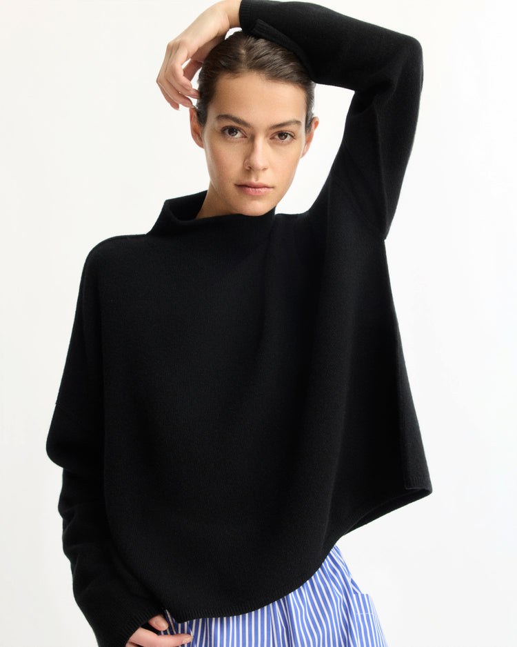 A woman with dark hair pulled back models the Arlo Mott Penn Roll Neck Top in Black, paired with blue-and-white striped pants—offering chic inspiration for spring layering against a plain white background.