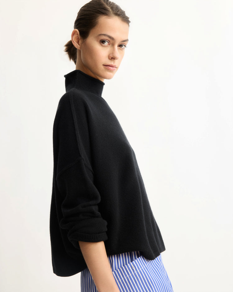 A woman with brown hair tied back wears the Arlo Mott Penn Roll Neck Top in Black with sleeves rolled up and blue-and-white striped pants, standing against a plain light background—an ideal look for effortless spring layering.