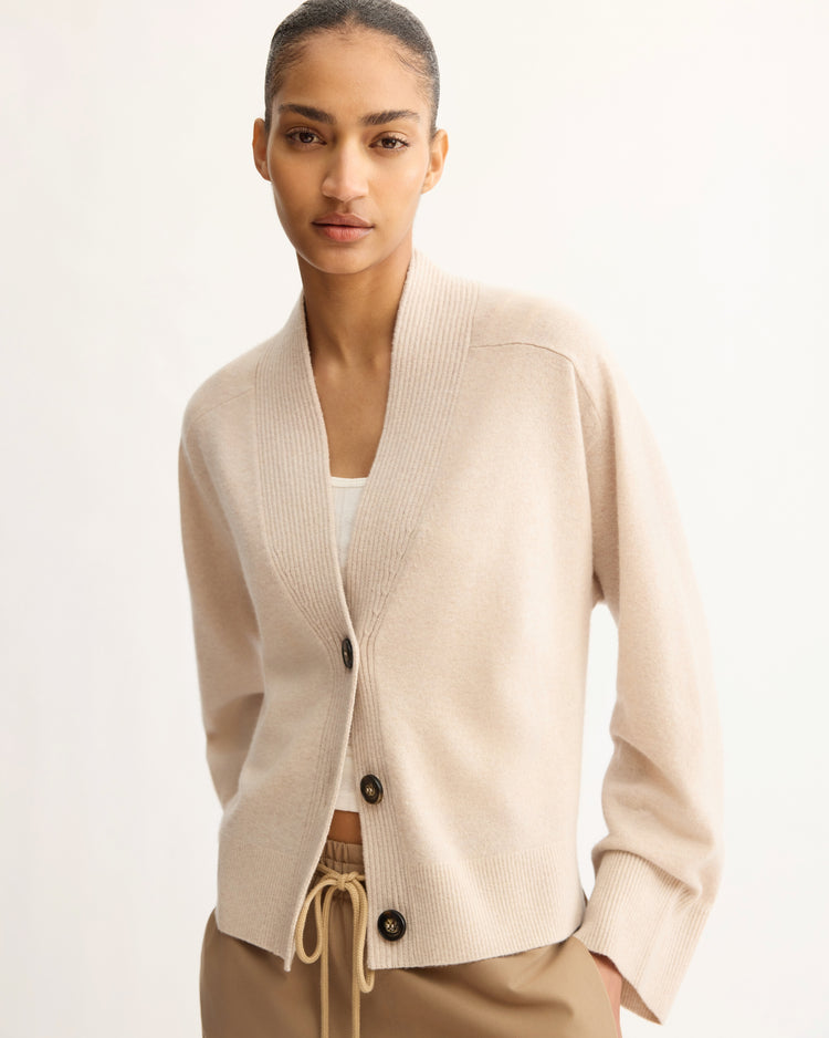 A woman with neatly pulled-back hair wears the Arlo Mott Wooster Cardigan in Beige over a white top and tan drawstring pants, standing against a plain light background.