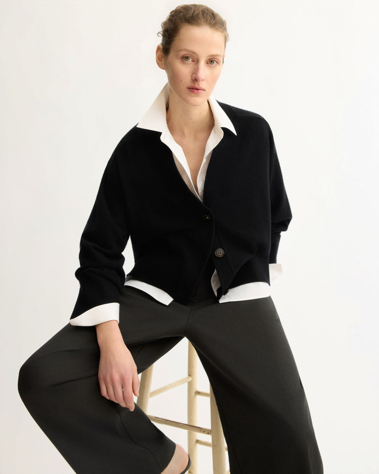 A person with light skin and tied-back hair sits on a stool, wearing the Arlo Mott Wooster Cardigan in Black over a white collared shirt and dark wide-leg pants against a plain light background.