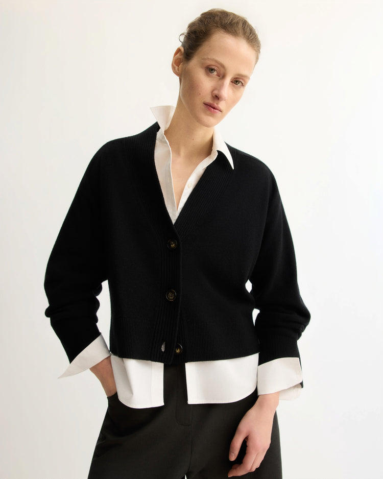 A person with light skin and tied-back hair stands in a relaxed pose against a plain light background, wearing the Arlo Mott Wooster Cardigan in Black over a white collared shirt and dark pants.
