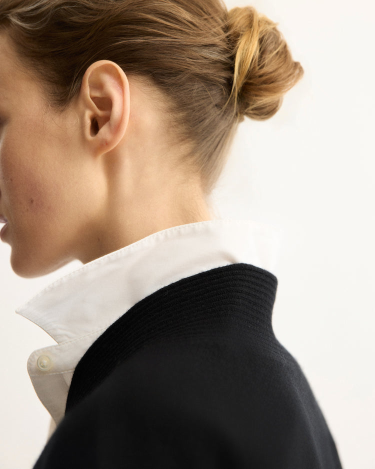 A close-up side view of a woman with light skin and brown hair in a bun, wearing the Arlo Mott Wooster Cardigan in Black over a white collared shirt, collar turned up, set against a neutral background.