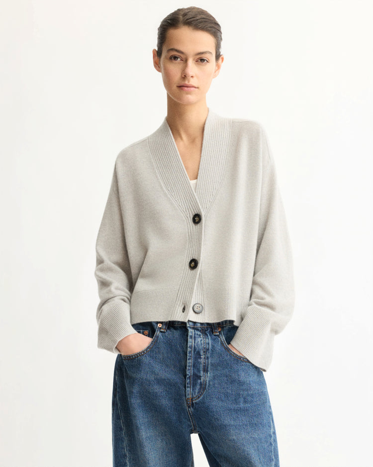 A person wears the Arlo Mott Wooster Cardigan in Grey—featuring large buttons—over a white top with high-waisted blue jeans, posing hands-in-pockets against a plain white background.