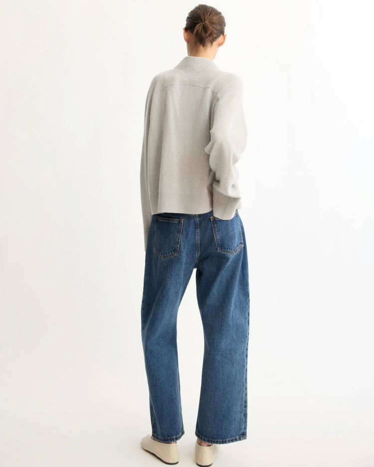 A person with their hair in a low bun faces away from the camera, wearing the Arlo Mott Wooster Cardigan in Grey, loose blue jeans, and white shoes against a plain white background.