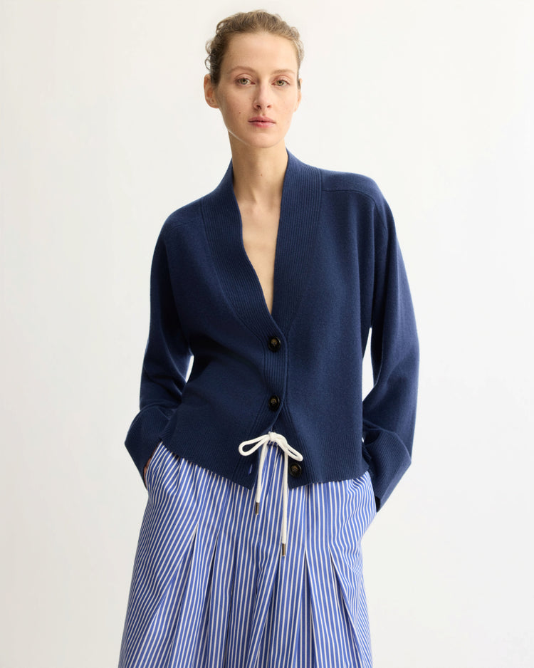 A person wears the Arlo Mott Wooster Cardigan in Navy with a white drawstring waist and a blue-and-white striped skirt, standing against a plain white background.