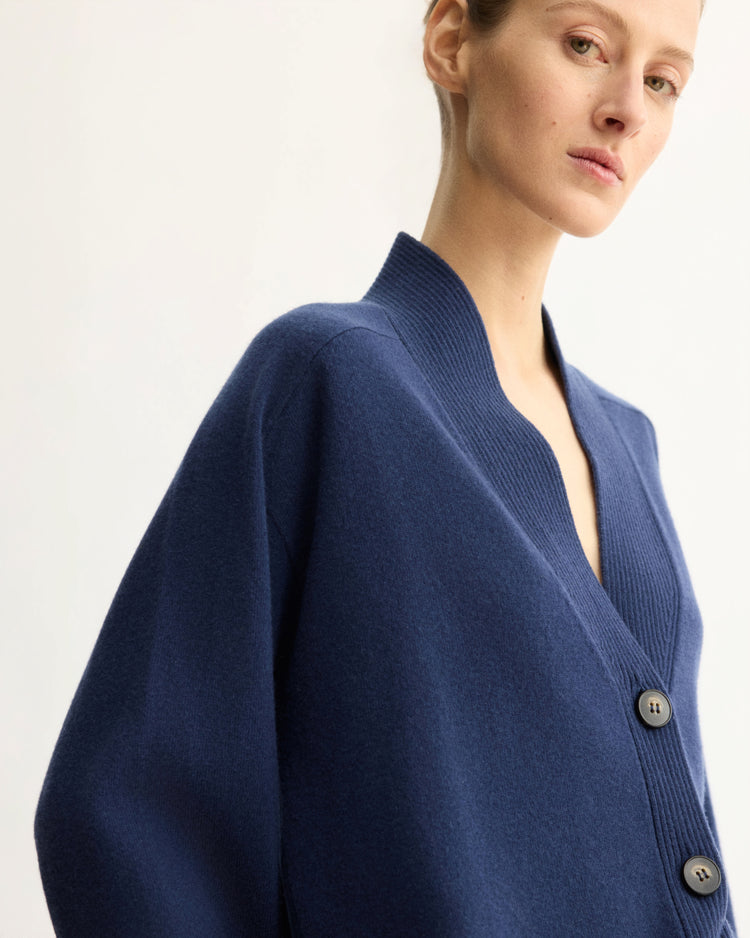 A person wears the Arlo Mott Wooster Cardigan in Navy—an oversized wool design with large black buttons—standing against a plain light background, gazing slightly to the side with a neutral expression.