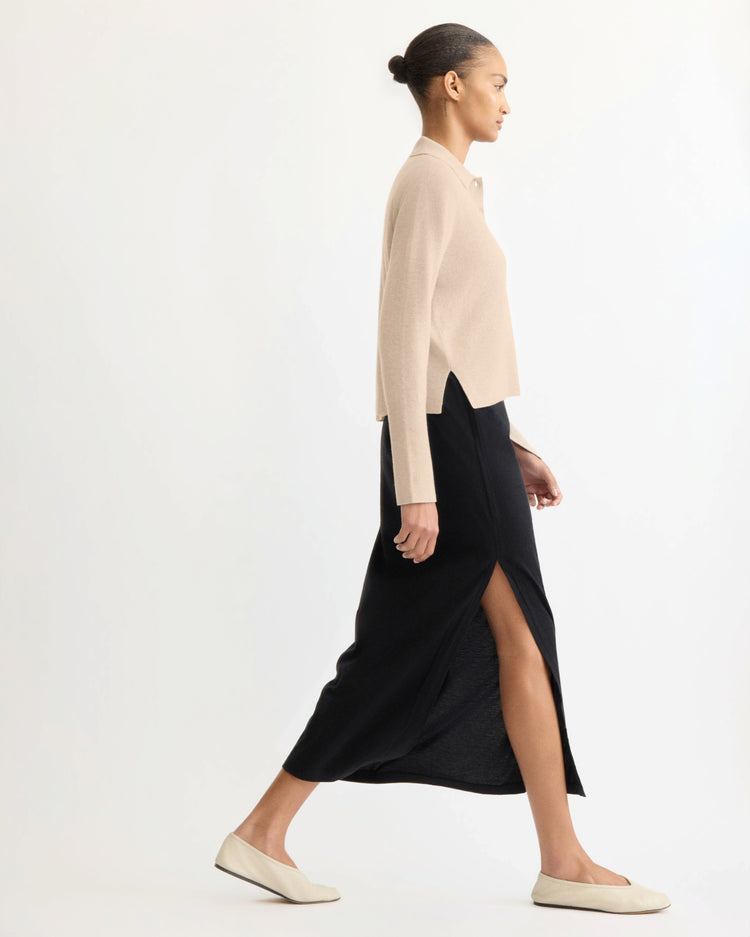 A woman with her hair in a bun walks in profile, wearing the Arlo Mott Berry Polo Long Sleeve in Beige, a black skirt with a side slit, and cream flats against a plain light background.