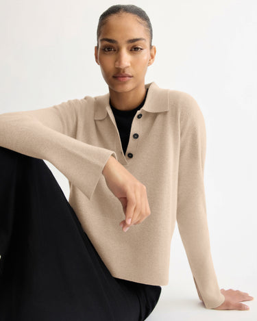 A person with neatly tied-back hair wears the Arlo Mott Berry Polo Long Sleeve in Beige with real horn buttons and black pants, seated on the floor against a plain white background.