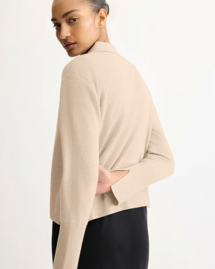 A person with neatly styled hair wears the Arlo Mott Berry Polo Long Sleeve in Beige and a black skirt, standing sideways and looking over their shoulder against a plain light background.