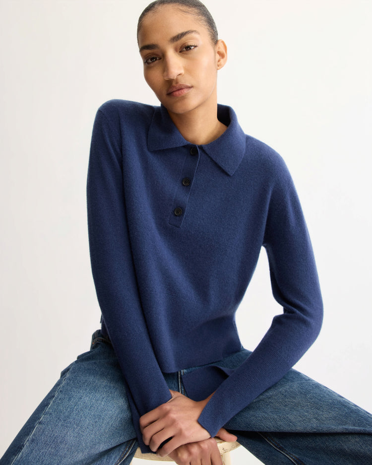 A person in the Arlo Mott Berry Polo Long Sleeve in Navy and blue jeans sits on a stool against a plain light background, confidently looking at the camera.