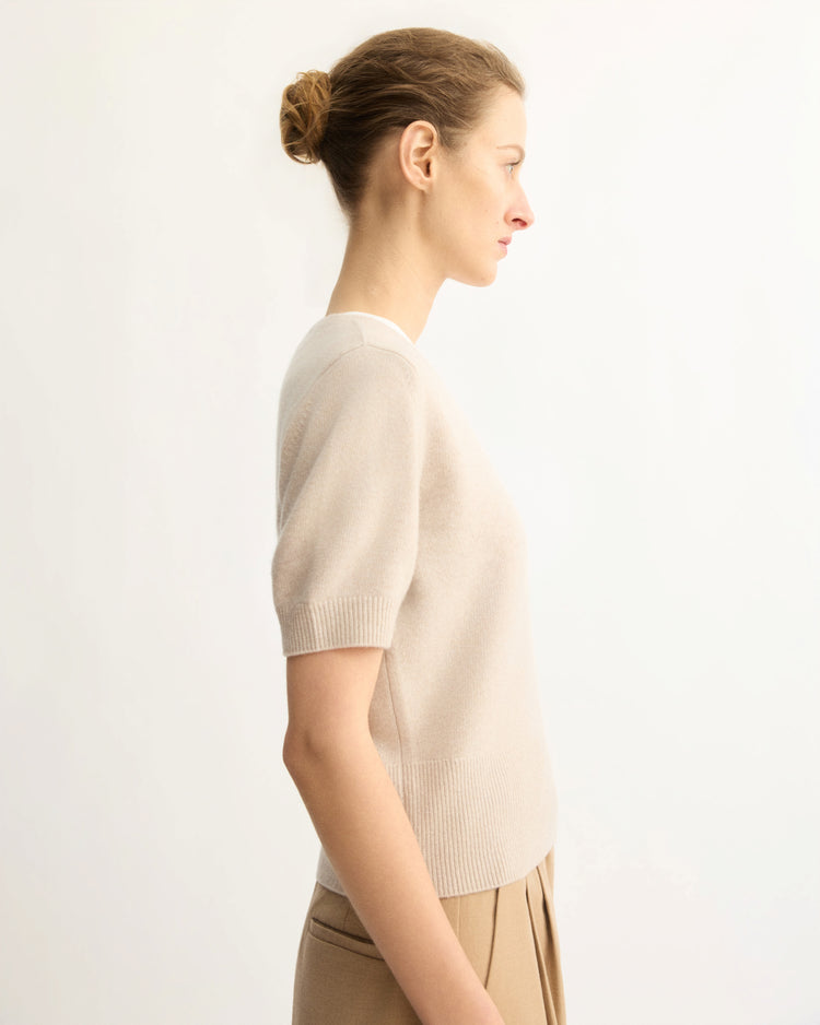 A woman with light skin and brown hair in a bun stands in profile against a plain light background, wearing the Arlo Mott Clarkson Short Sleeve Crewneck in Beige and tan pleated pants—a perfect seasonal staple.