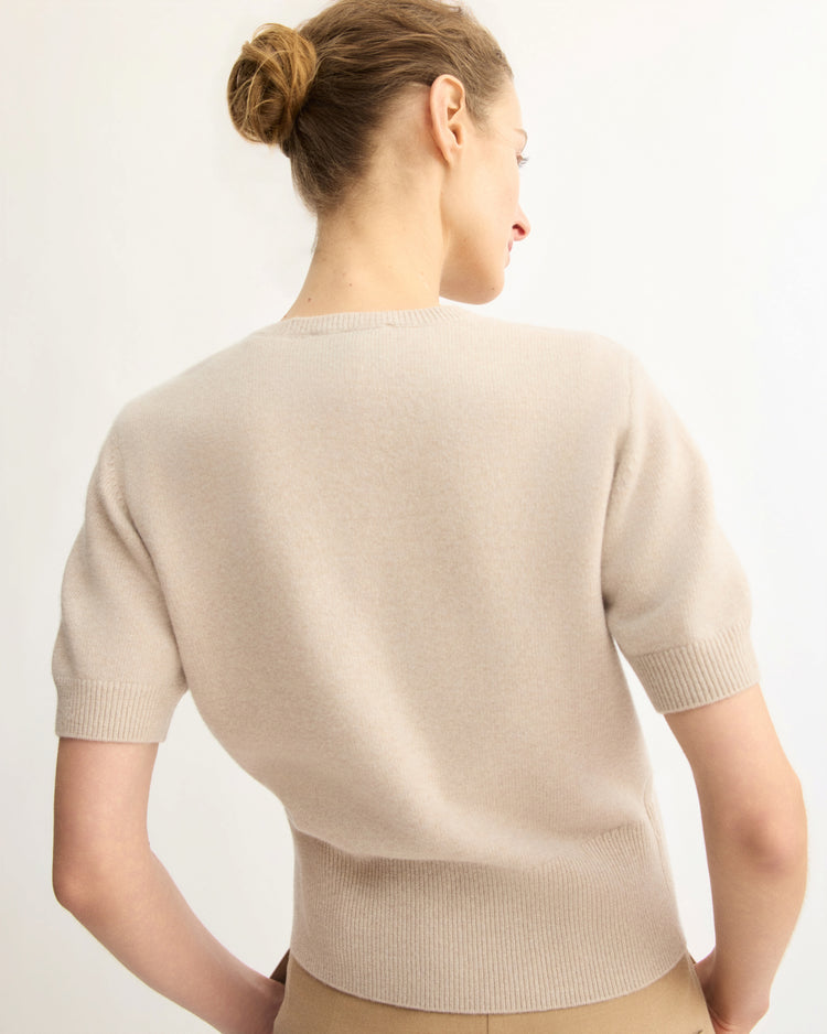 A woman with light brown hair in a bun is seen from the back wearing the Arlo Mott Clarkson Short Sleeve Crewneck in Beige and tan pants, standing against a plain, light-colored background.