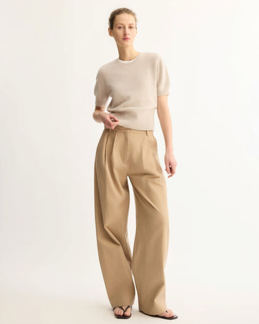 A person stands against a light background wearing the Arlo Mott Clarkson Short Sleeve Crewneck in Beige, tucked into high-waisted, wide-leg tan trousers with black sandals—a relaxed look with one hand in a pocket.