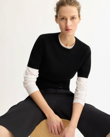 A person with light skin and short, curly hair sits on a stool, wearing the Arlo Mott Clarkson Short Sleeve Crewneck in Black over a white long-sleeve shirt and black pants, looking at the camera against a plain light background.