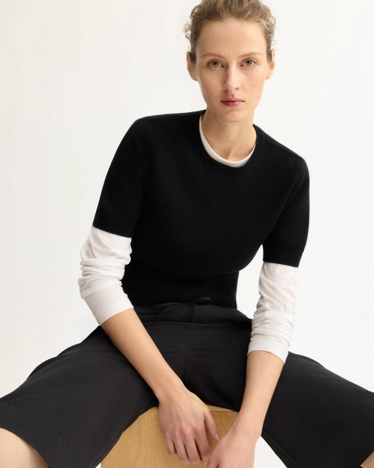 A person with light skin and short, curly hair sits on a stool, wearing the Arlo Mott Clarkson Short Sleeve Crewneck in Black over a white long-sleeve shirt and black pants, looking at the camera against a plain light background.