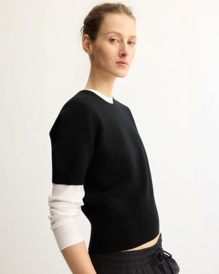A person with light skin and pulled-back hair stands against a plain background, wearing the Arlo Mott Clarkson Short Sleeve Crewneck in Black over a white long-sleeve shirt and dark pants, looking slightly to the side with a neutral expression.