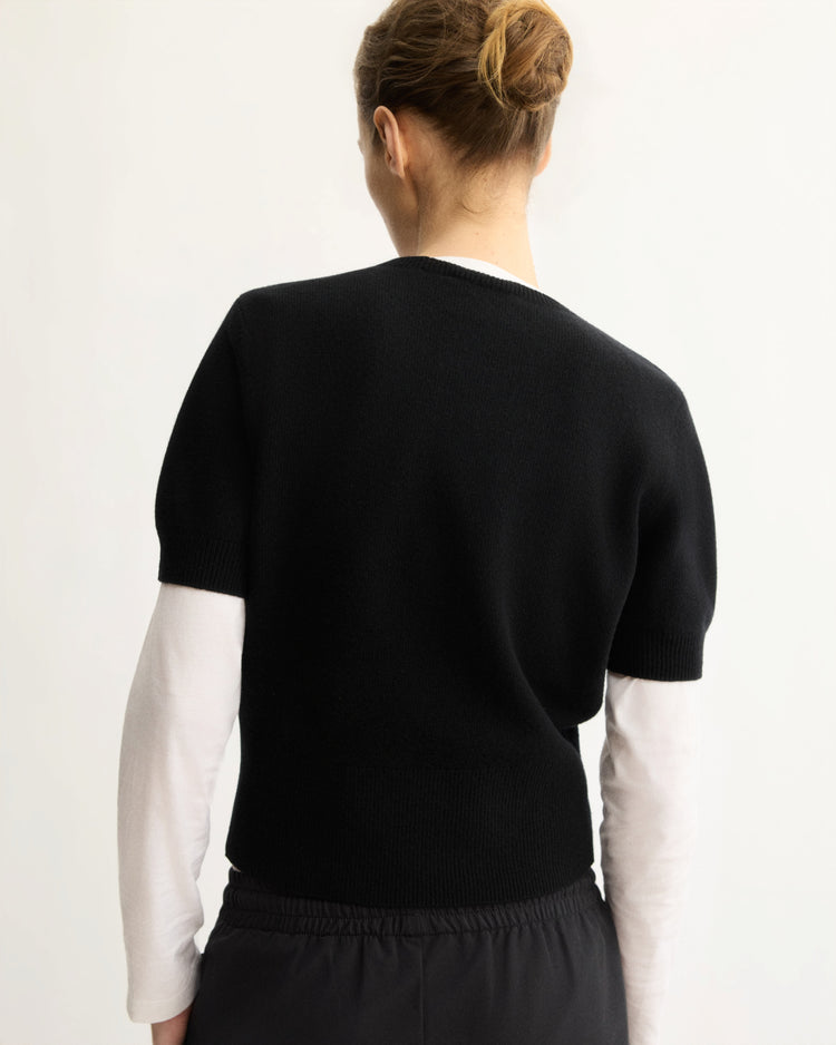 A light-skinned person with brown hair in a bun faces away, wearing Arlo Mott’s Clarkson Short Sleeve Crewneck in Black over a white long-sleeve shirt and black pants, standing against a plain light background.