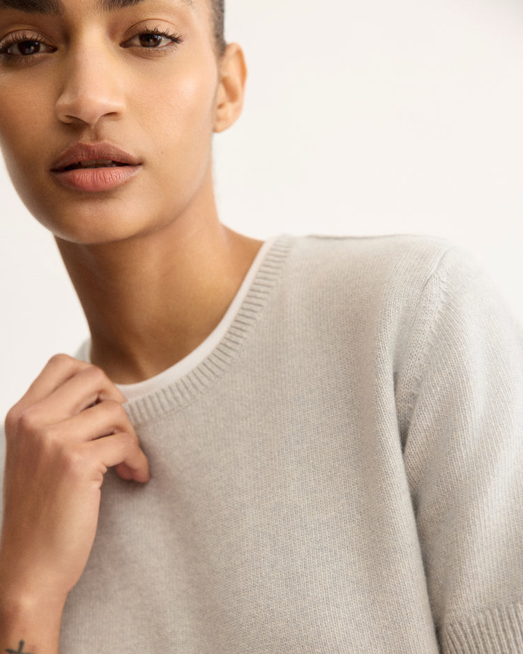 A person with short hair is shown from the shoulders up, wearing the Arlo Mott Clarkson Short Sleeve Crewneck in Grey, looking slightly to the side with a neutral expression against a softly lit background.