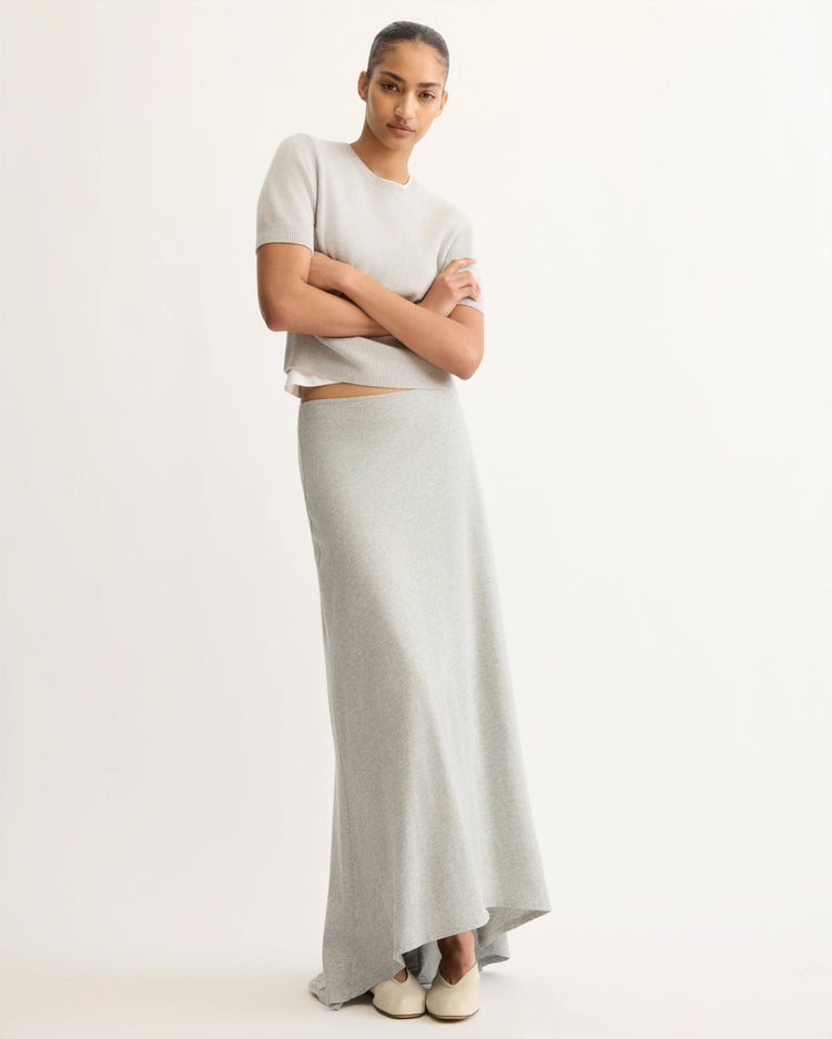 A woman stands against a plain background wearing the Arlo Mott Clarkson Short Sleeve Crewneck in grey, paired with a matching long skirt and white flat shoes. She crosses her arms and looks confidently at the camera.