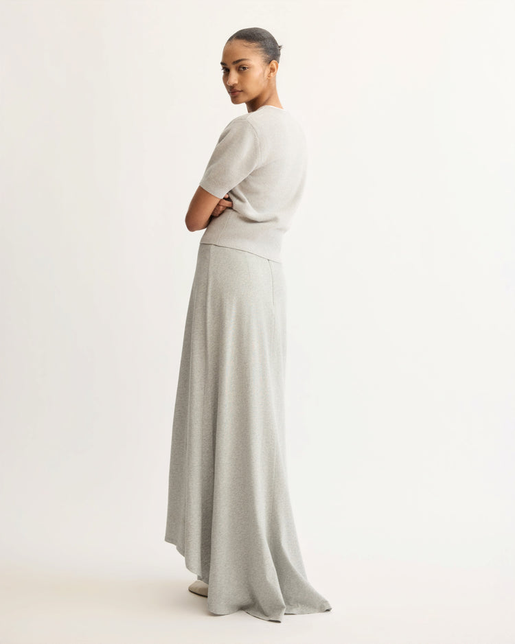 A woman with her hair in a bun stands sideways, looking over her shoulder. She wears a light gray short-sleeve sweater and the Arlo Mott Beekman Maxi Skirt in Grey, which drapes elegantly against a plain white background.