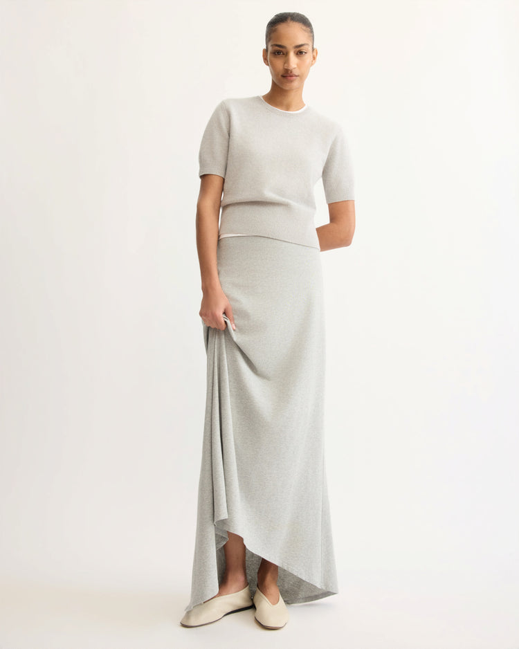 A woman poses against a light background, wearing the Arlo Mott Beekman Maxi Skirt in Grey with a short-sleeve, light gray cotton jersey top and beige slip-on shoes. She holds part of her skirt and looks at the camera.