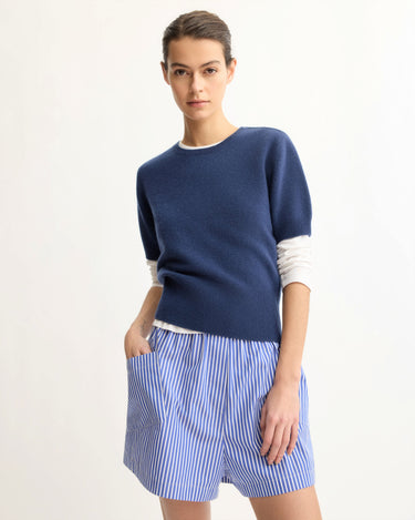 A woman stands against a plain light background wearing the Arlo Mott Clarkson Short Sleeve Crewneck in Navy over a white long-sleeved shirt and a blue-and-white vertically striped skirt, with one hand in her pocket.