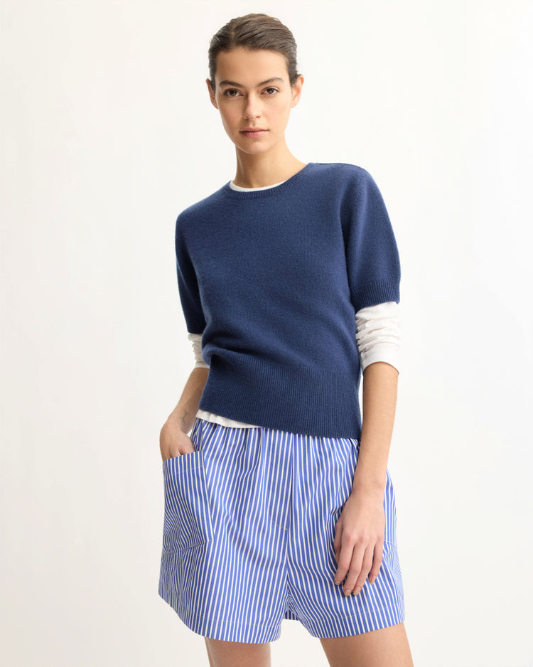 A woman stands against a plain light background wearing the Arlo Mott Clarkson Short Sleeve Crewneck in Navy over a white long-sleeved shirt and a blue-and-white vertically striped skirt, with one hand in her pocket.