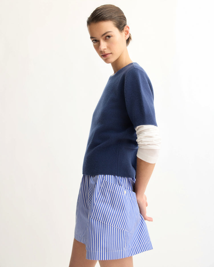 A woman in profile wears the Arlo Mott Clarkson Short Sleeve Crewneck in Navy over a white long-sleeve shirt and a blue-and-white striped skirt, posing against a plain background with a neutral expression.