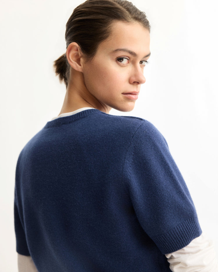 A woman with brown hair tied back looks over her shoulder at the camera, wearing the Arlo Mott Clarkson Short Sleeve Crewneck in Navy over a white shirt, set against a plain light background.