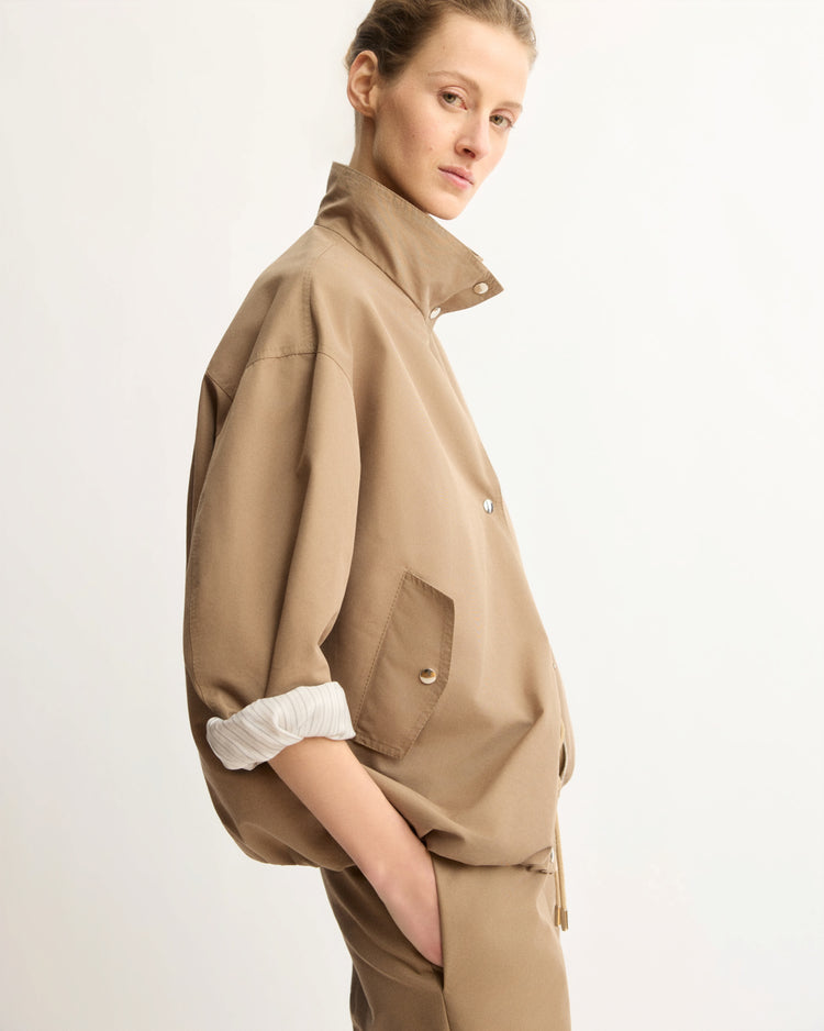A person with light hair pulled back wears the Arlo Mott Logan Tech Jacket in Khaki with rolled-up sleeves and matching pants, standing with one hand in their pocket against a plain background.