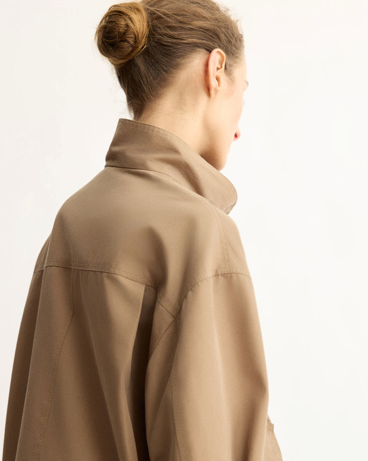 A person with light brown hair in a bun is seen from behind, wearing the Arlo Mott Logan Tech Jacket in Khaki, standing against a plain light background.