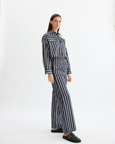A woman stands against a white background, wearing Arlo Mott's Hudson Pull-on Pants in Navy Stripe—wide-leg, crafted from soft Portuguese cotton—and a matching striped button-up shirt, paired with black slip-on shoes.